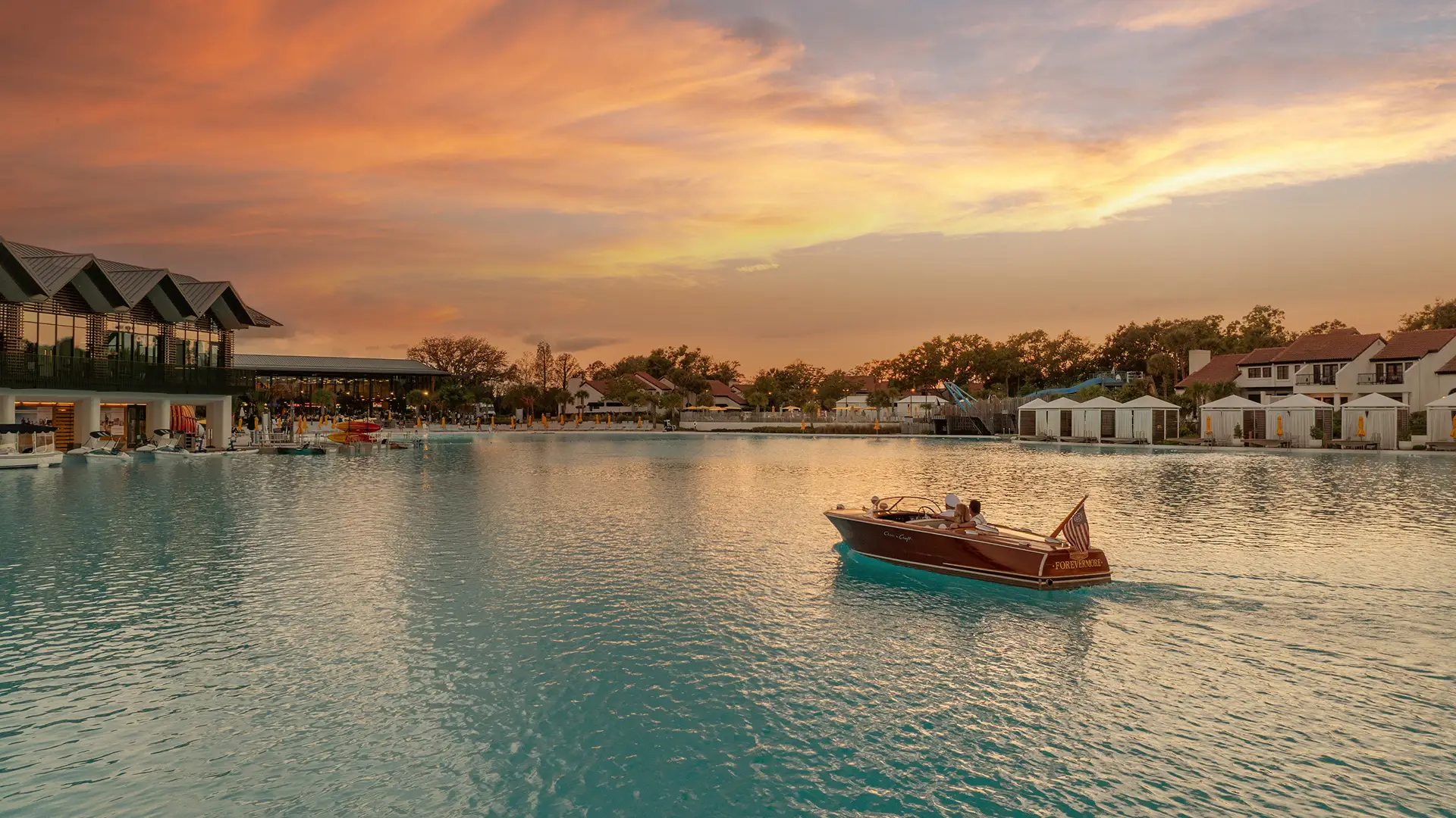 Couple riding a wooden boat on large blue lagoon at sunset with buildings and cabanas