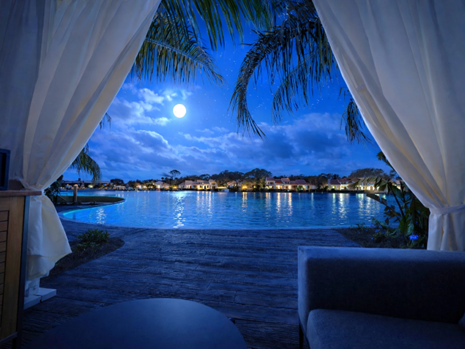 View of a moonlit lake with houses and palm trees, seen from a cabana with curtains and outdoor seating.