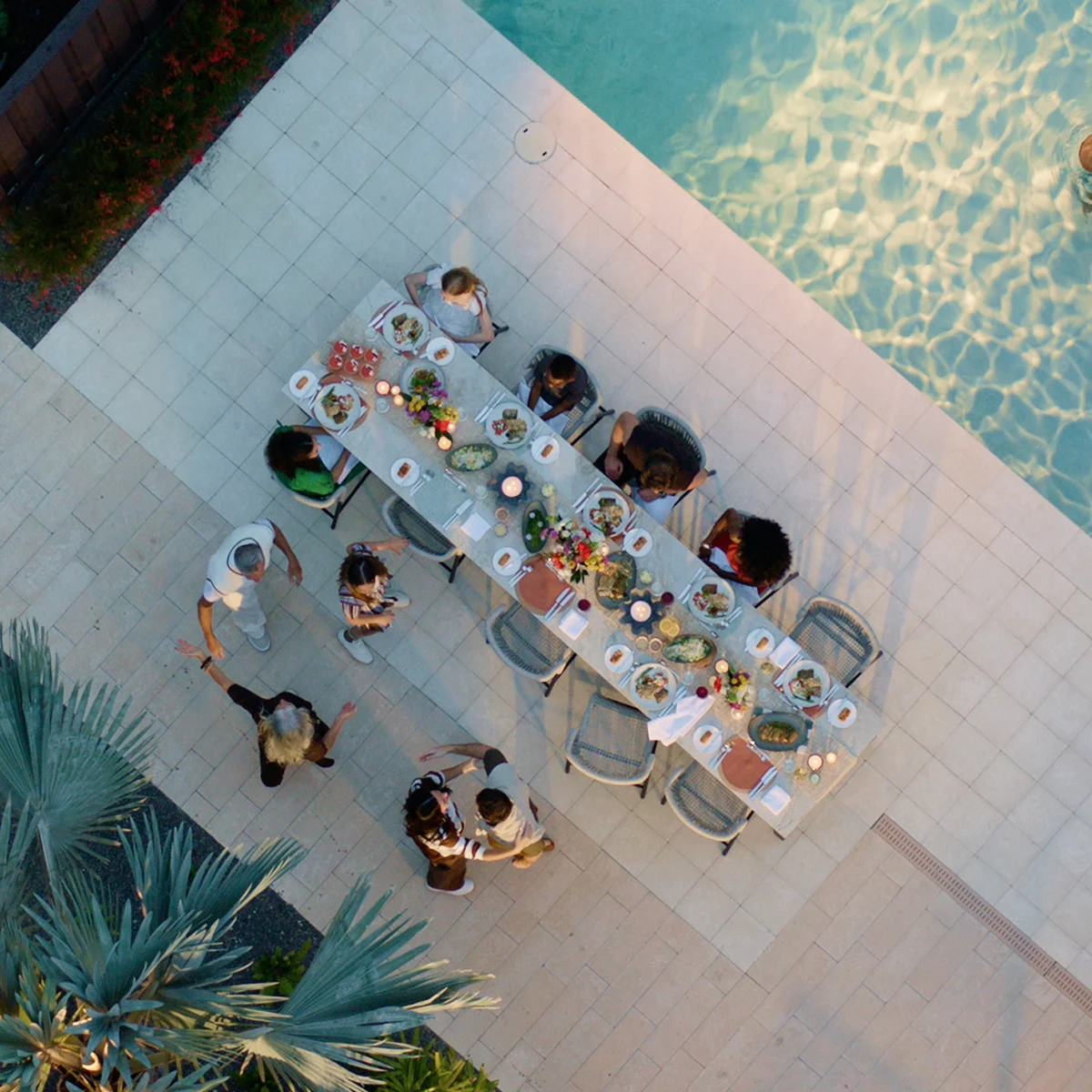 Overhead view of people dining outdoors by a pool with a long table set with meals and candles.