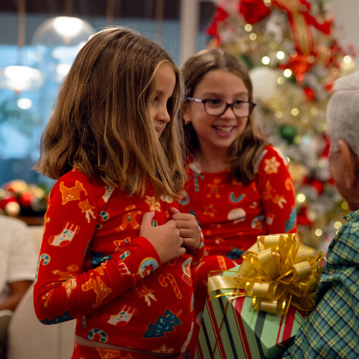 Two girls in red holiday pajamas receive a wrapped gift with a large gold bow near a Christmas tree.