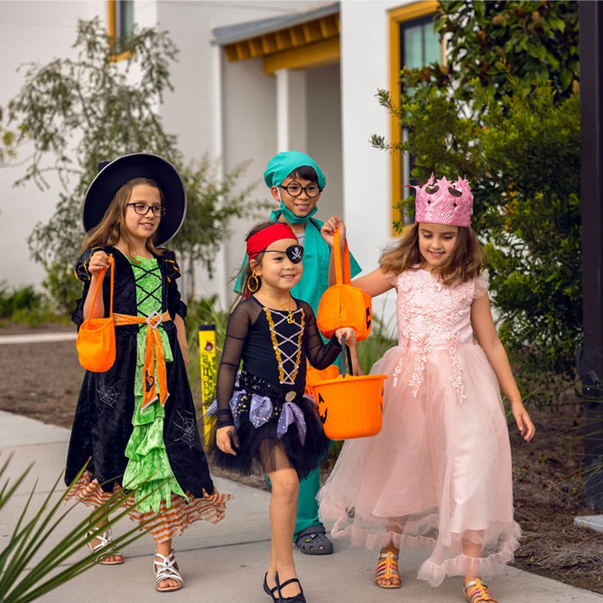 Four children in Halloween costumes, including witch, pirate, doctor, and princess, carrying orange treat bags.
