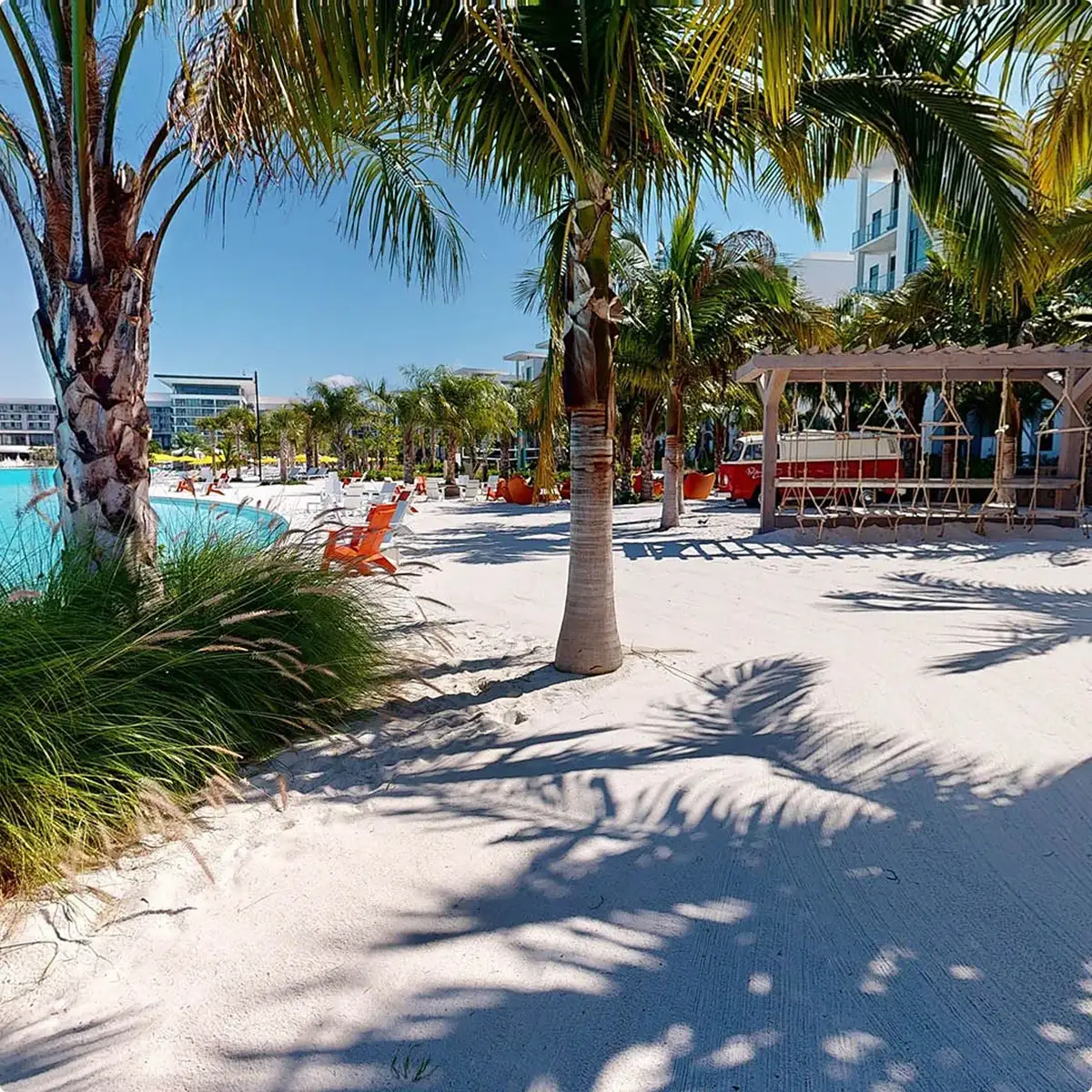 Palm trees casting shadows on white sand near a pool with orange chairs and a swing structure.
