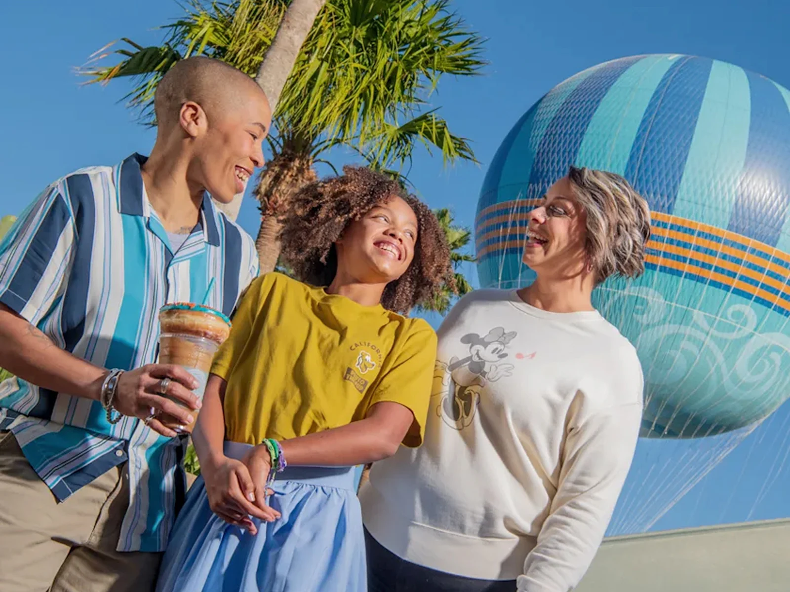 Three people smiling together outdoors with palm trees and a large blue hot air balloon behind them.