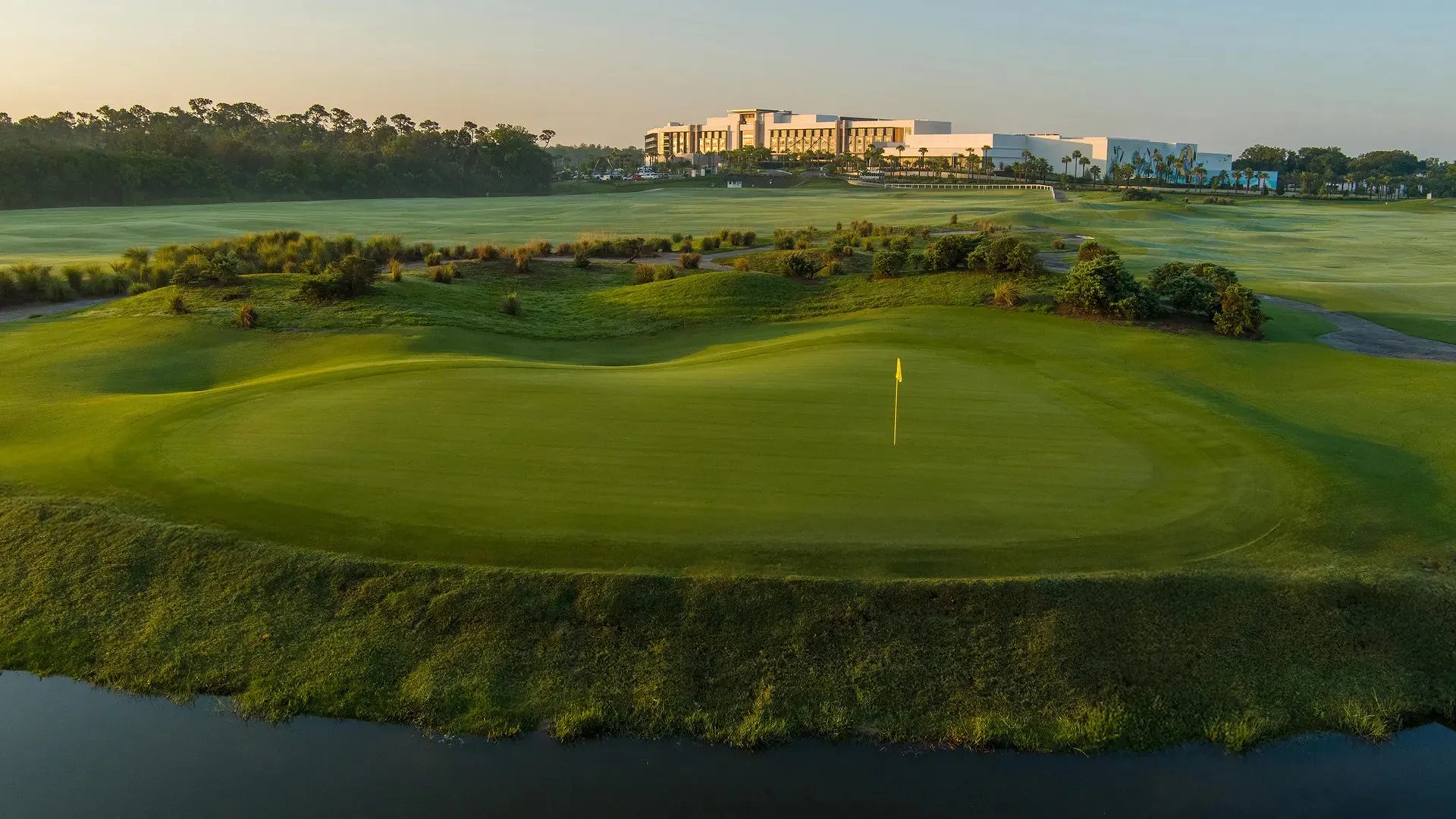 Golf green with yellow flag near water and bushes, with large building and trees in background at sunrise.
