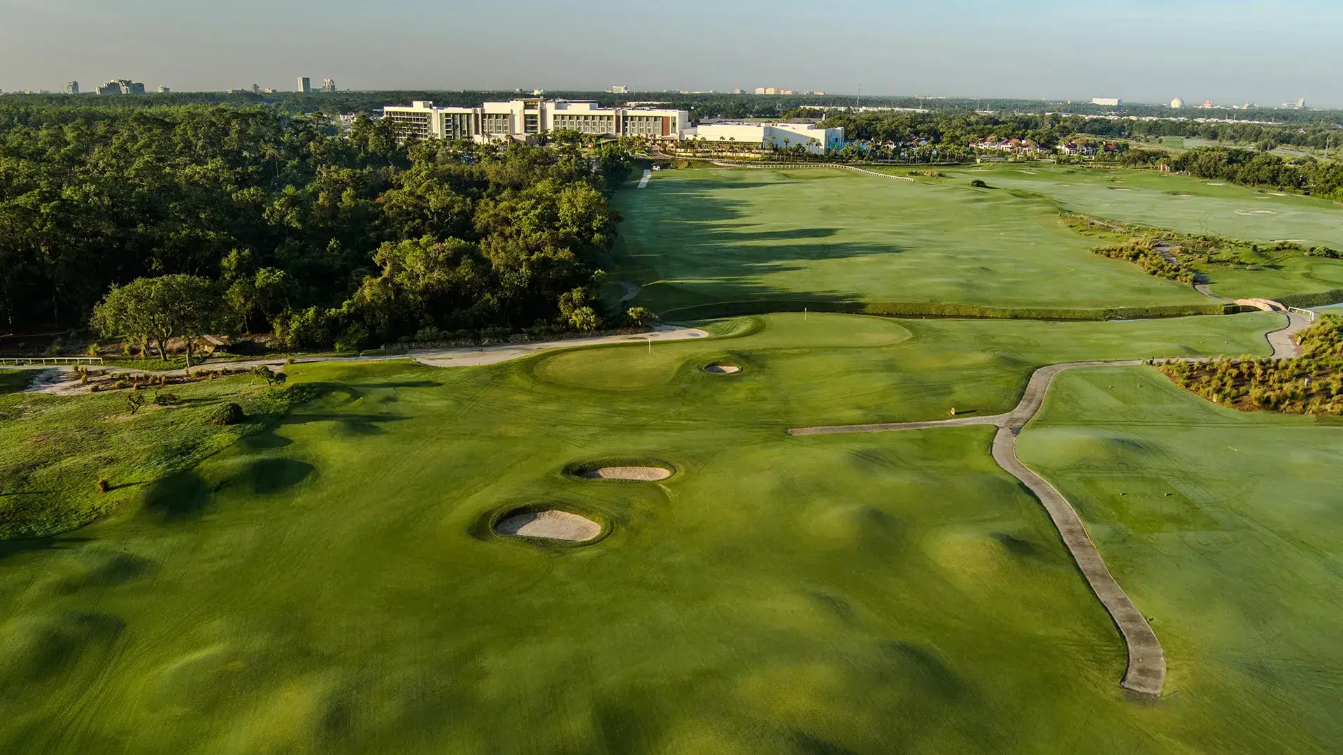 Aerial view of a golf course with sand bunkers and a path leading to a building surrounded by trees.