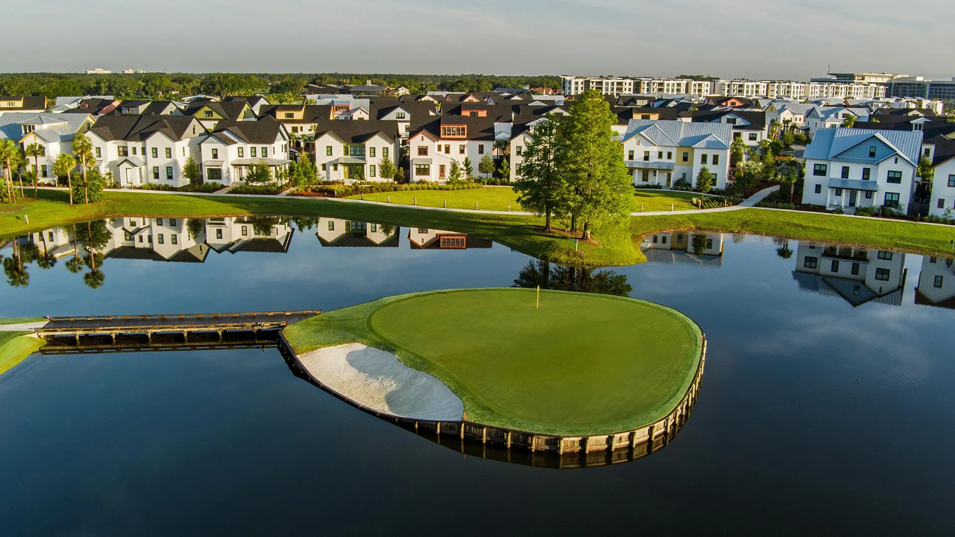 A golf green with sand trap on a water-surrounded island in front of suburban houses reflected in the water.