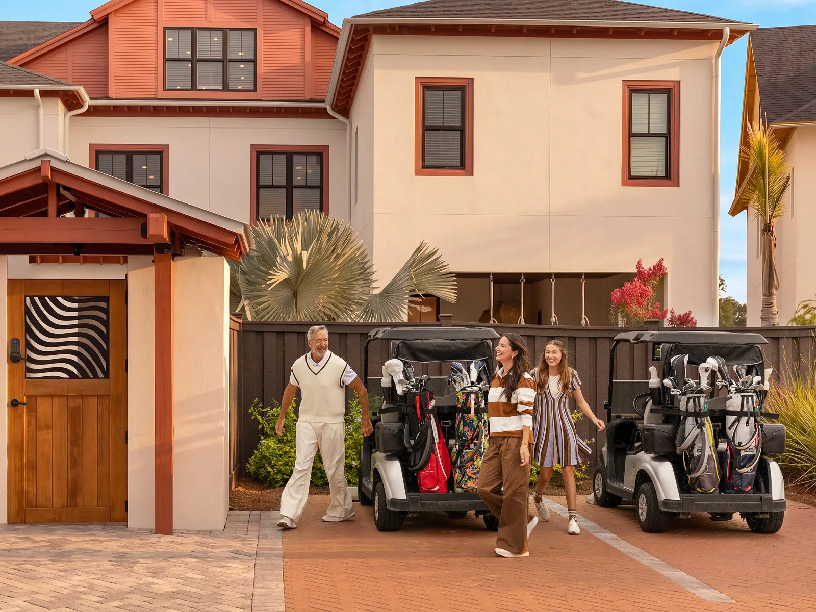 Man and two women walking past golf carts with bags outside a large modern house.