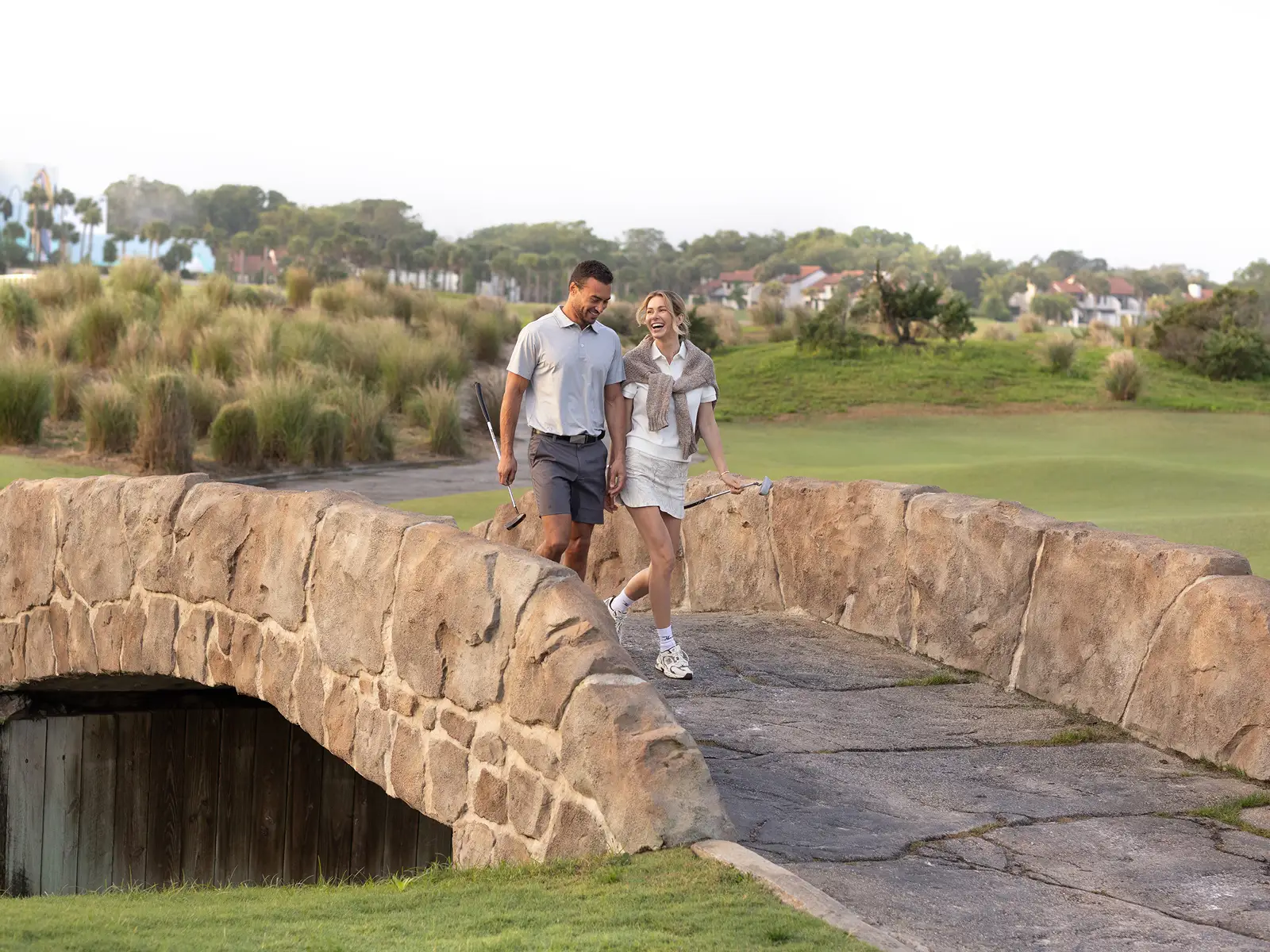 Man and woman walking on stone bridge at golf course holding golf clubs and smiling.