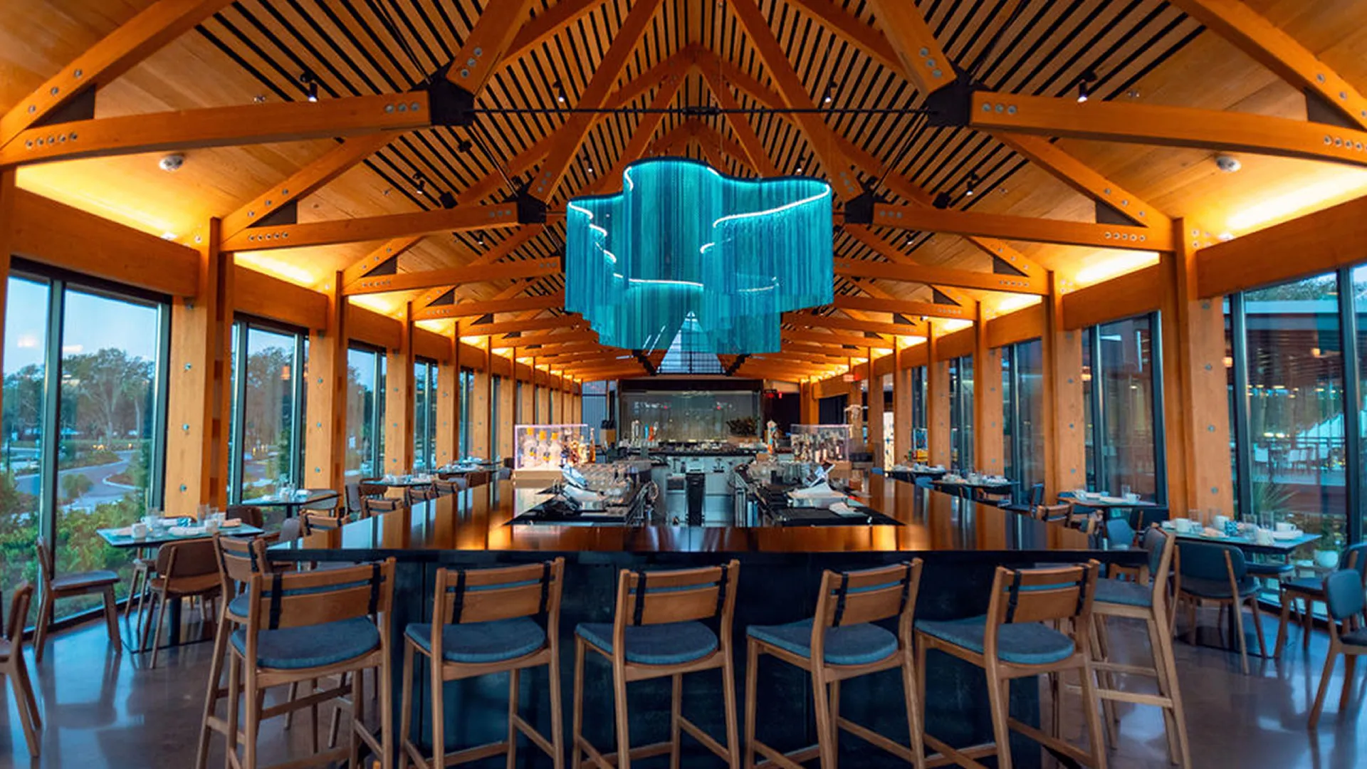 Modern restaurant interior with wooden beams, large windows, central bar, and a blue chandelier.