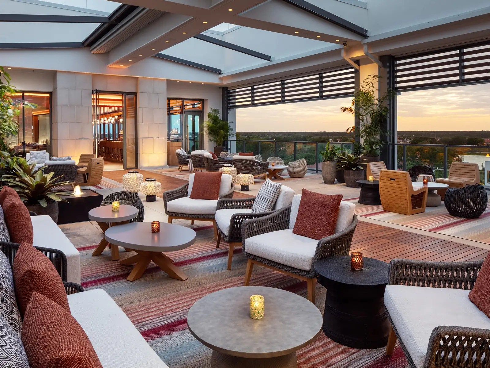 Outdoor rooftop lounge with cushioned chairs, tables, plants, and a view of the sunset over trees.