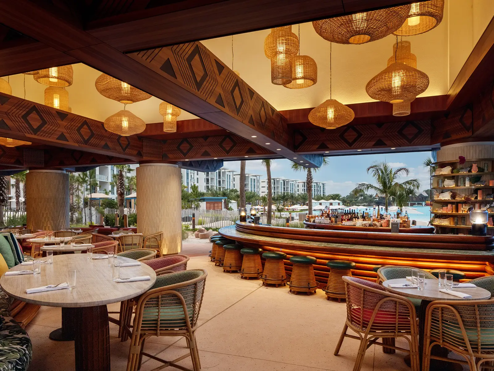 Open-air bar and dining area with wicker chairs, wooden tables, hanging lamps, and a pool view.