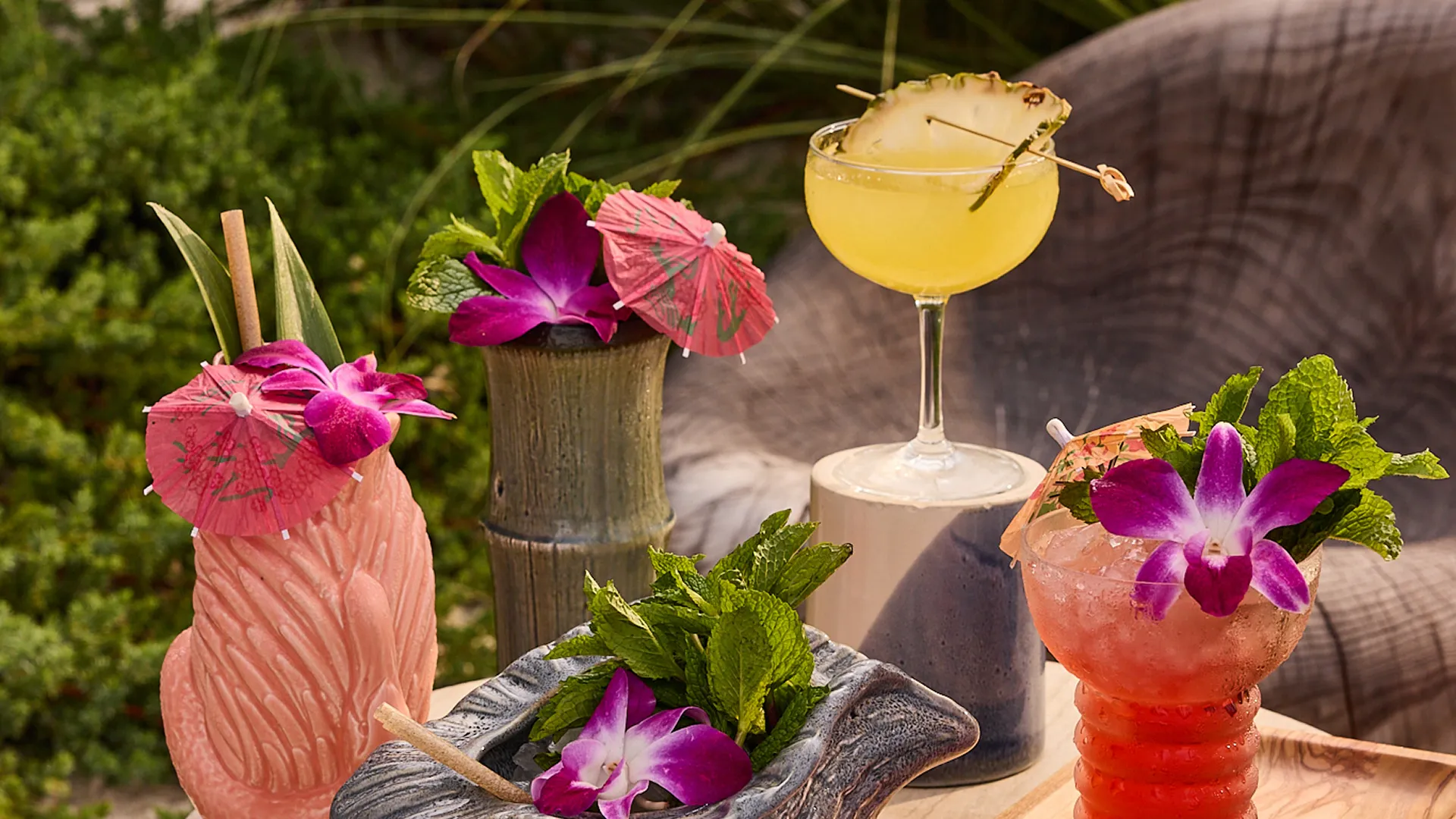 Four colorful tropical cocktails garnished with flowers, mint, umbrellas, and pineapple slice outdoors.