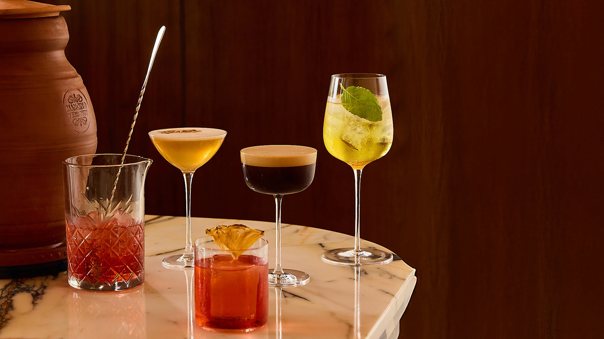 Five cocktails on a marble table, including a red drink with dried pineapple and a yellow drink with a mint leaf.
