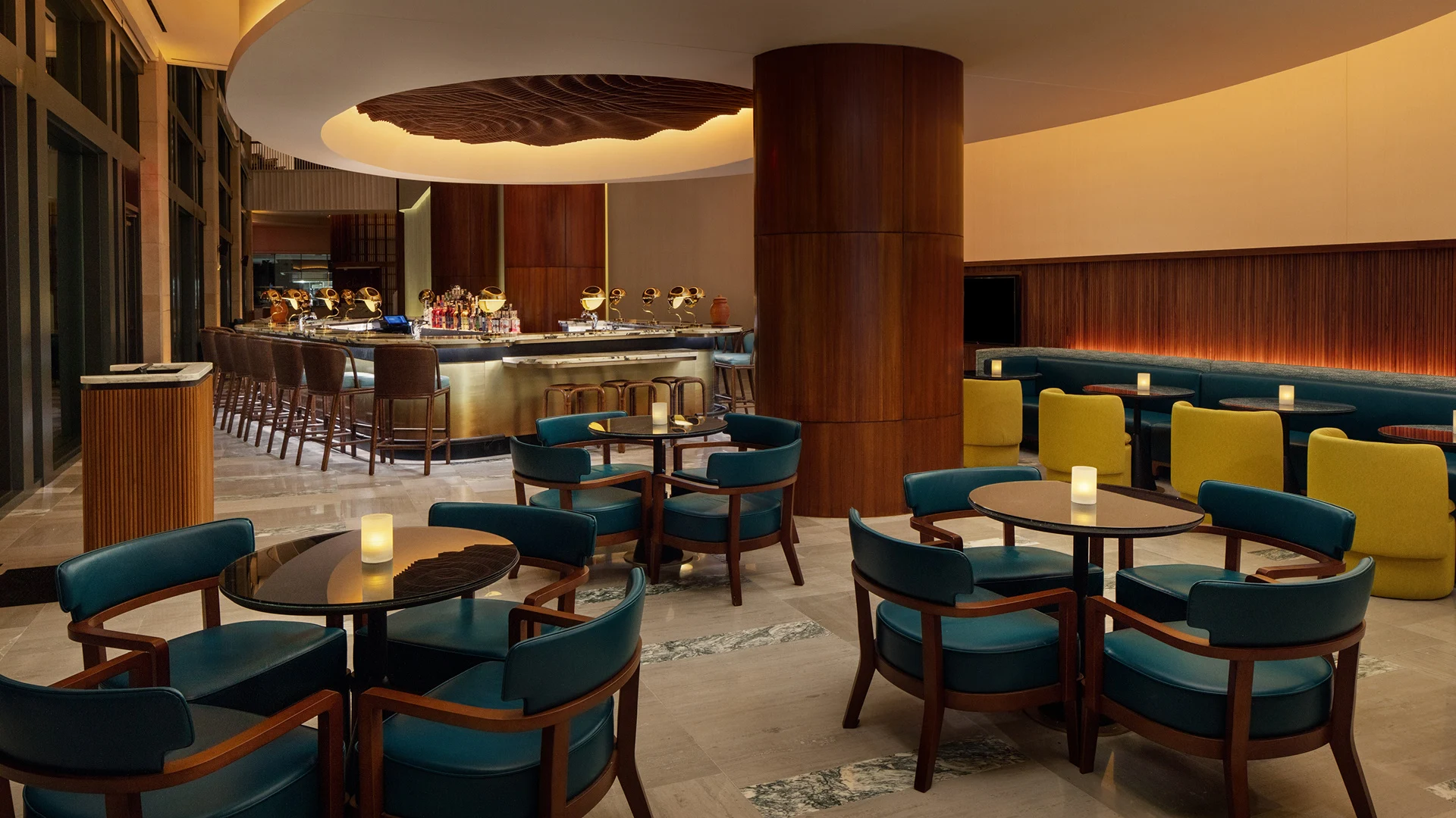 Modern hotel bar with teal chairs, round tables, a curved bar with brown stools, and ambient lighting.