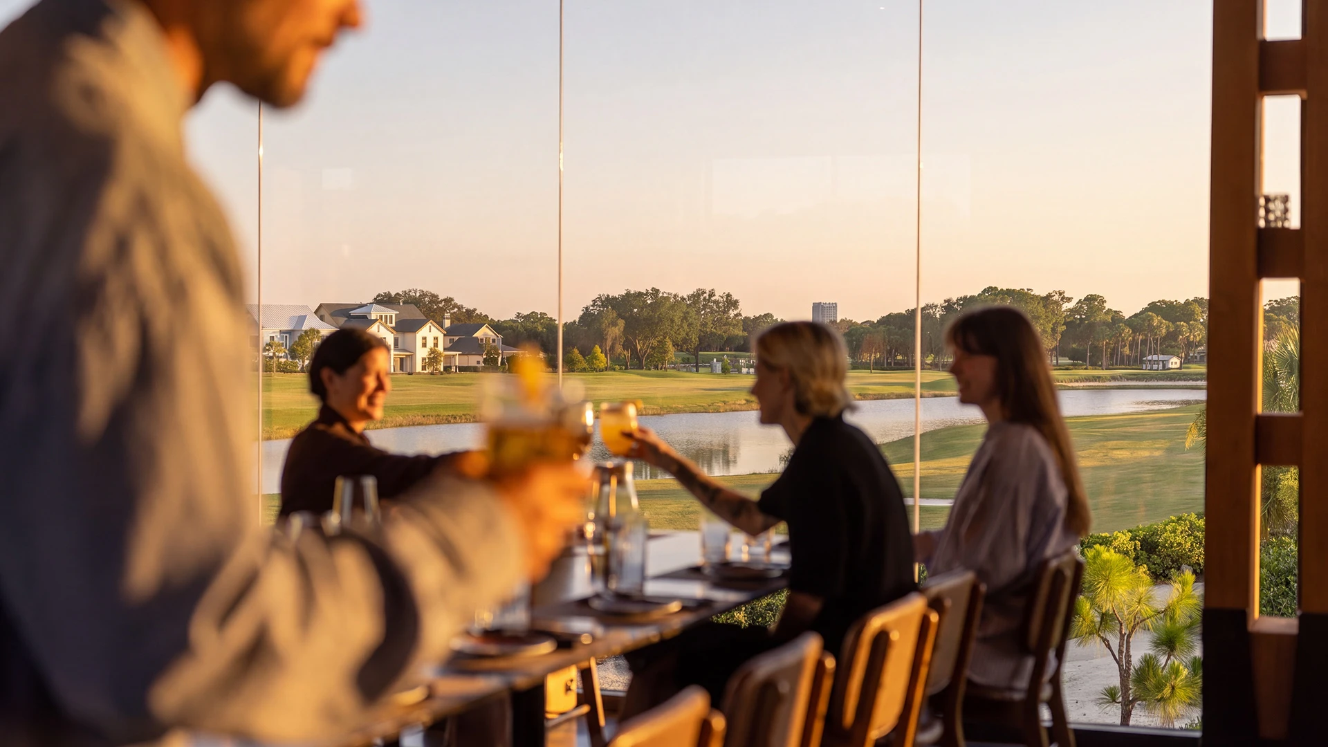 People sitting at a table inside raising drinks with a river and houses visible outside large windows.