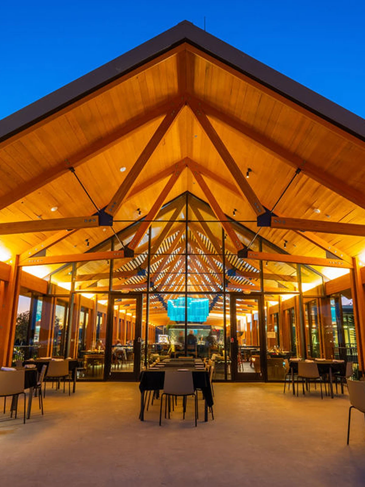 Empty outdoor dining area with tables and chairs under a wooden roof and glass walls at dusk.