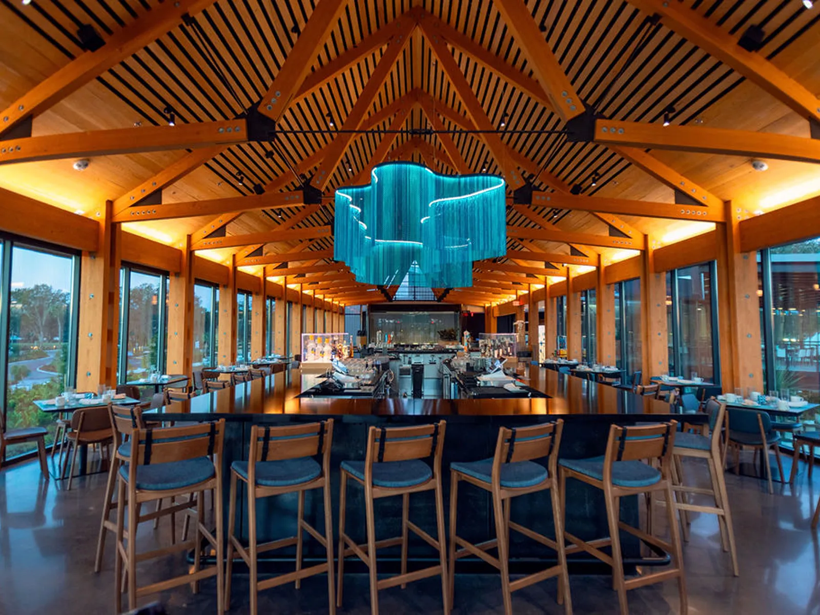 Modern restaurant interior with wooden beams, large windows, bar with high chairs, and blue chandelier.
