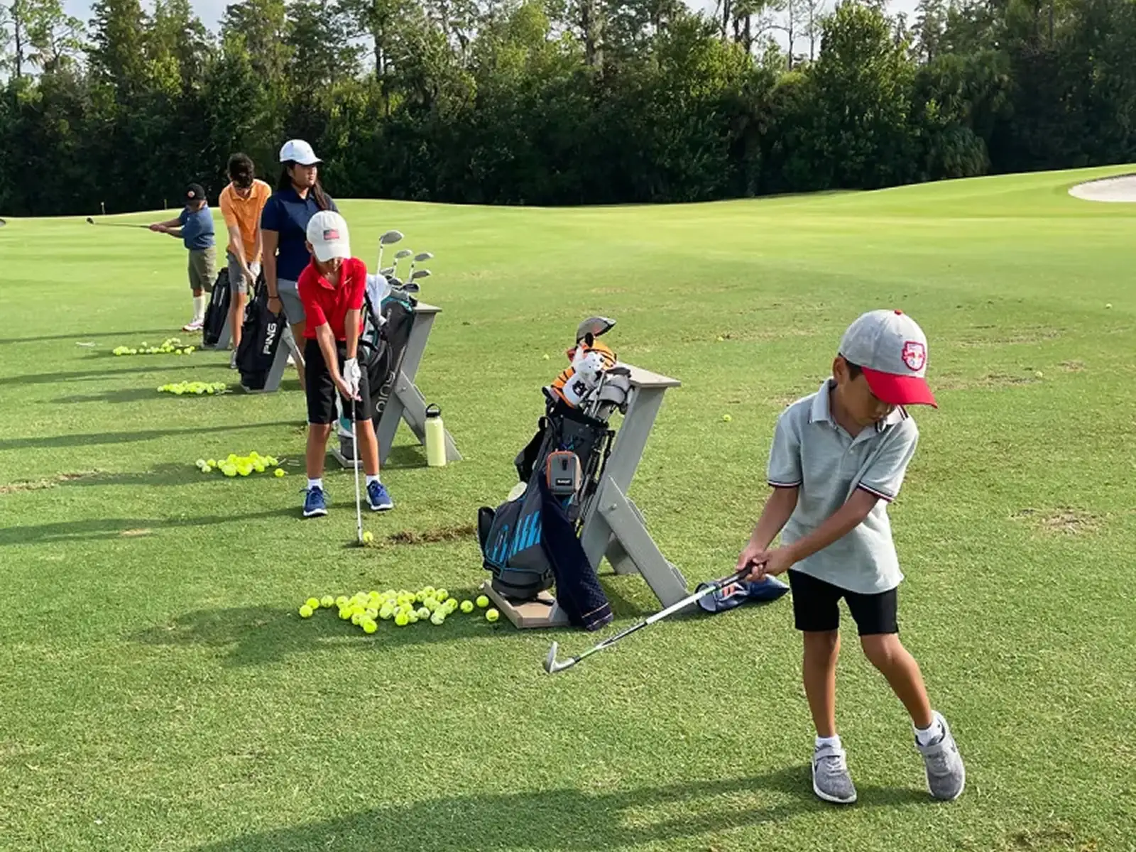 Children practicing golf swings on a grassy driving range with golf bags and balls lined up.