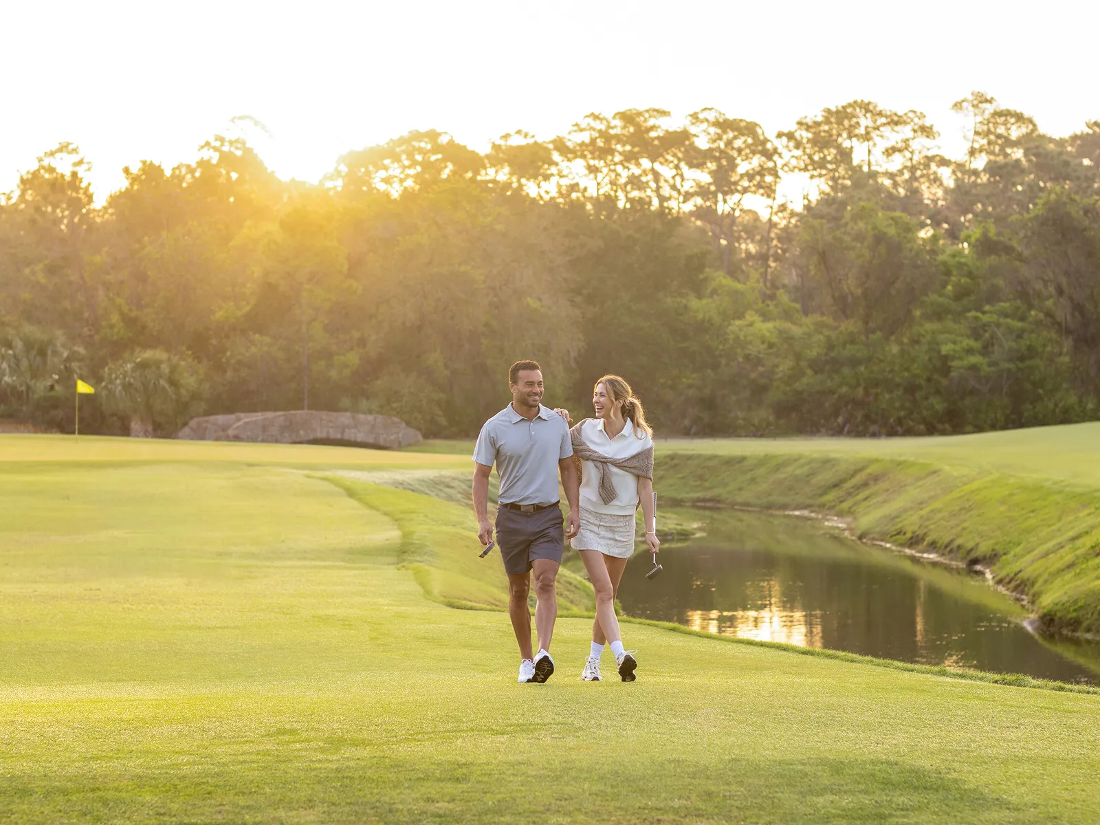 Man and woman walking on golf course near water, holding putters, with sunset and trees in background.