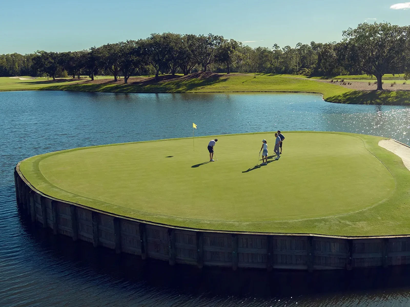 Three golfers on a green surrounded by water with a yellow flag on a sunny day.