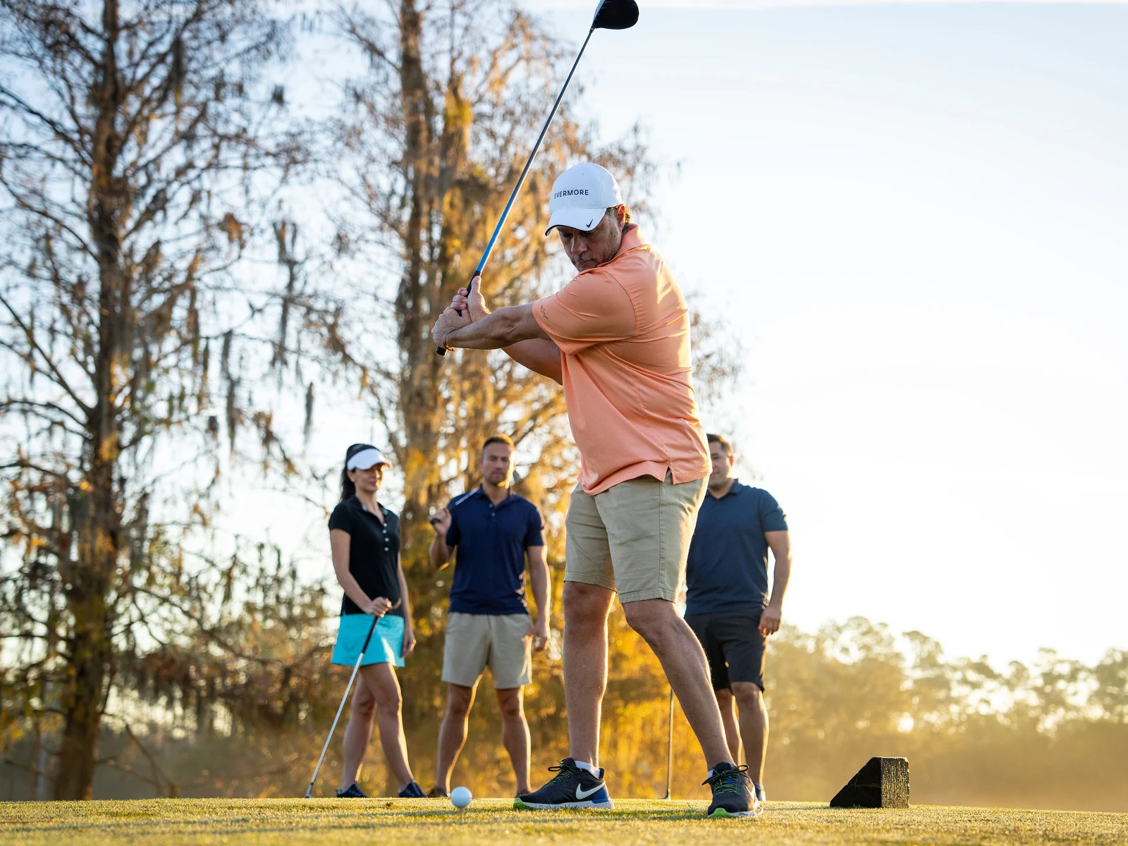 Man in orange shirt swinging golf club while three people watch on a sunny golf course.