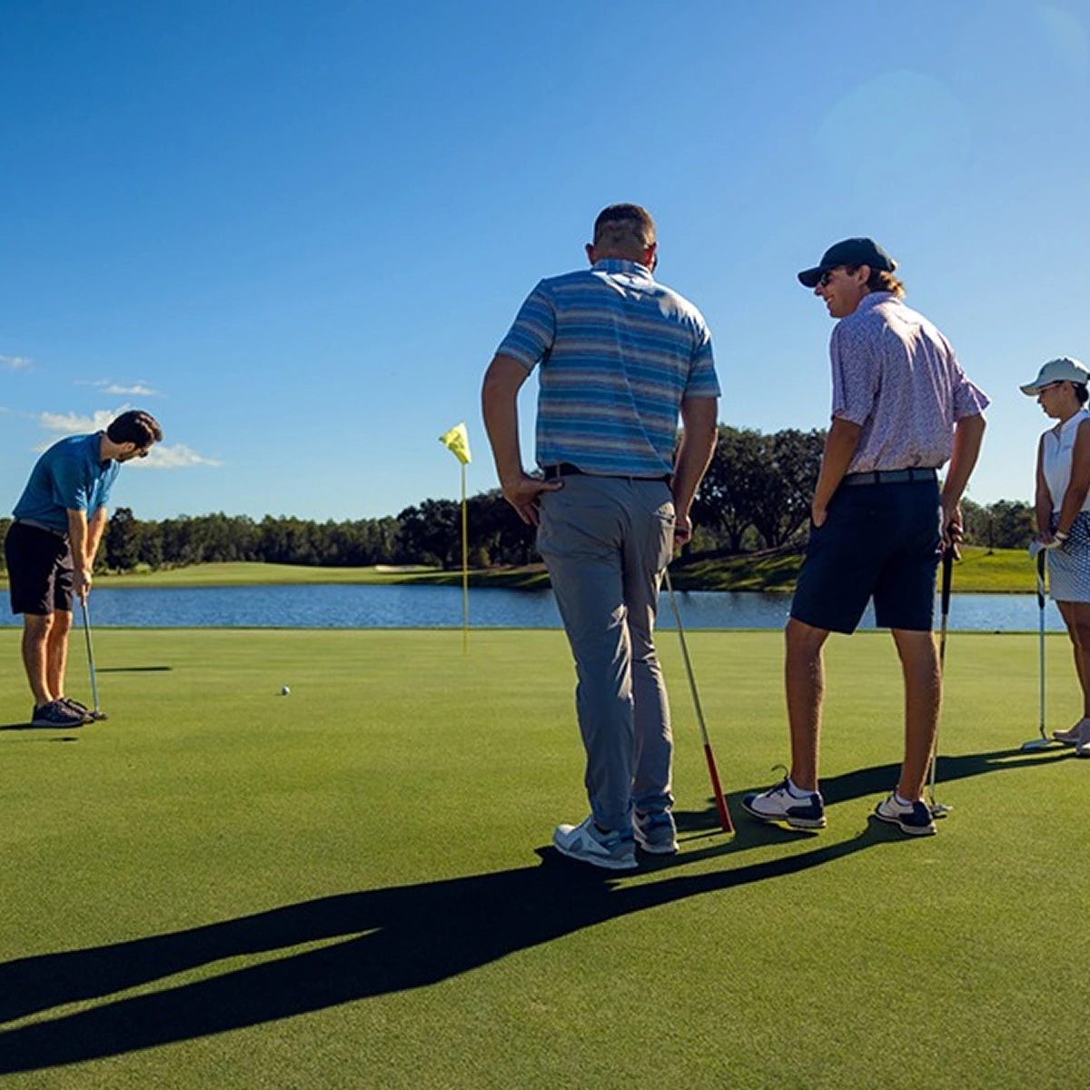 Four people playing golf on a green near a water body under clear blue sky.