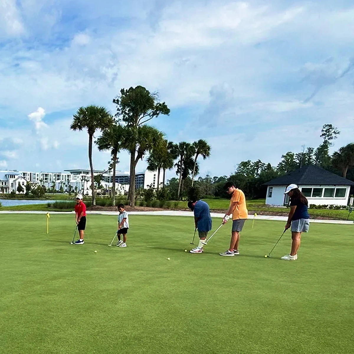 Five young golfers on a putting green aiming golf balls towards holes with flags in a sunny outdoor course.