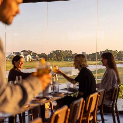 People sitting at a restaurant table by large windows, toasting drinks with a lake and houses outside.