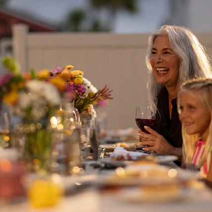 Smiling older woman holding wine glass at a dinner table with a young girl and flowers.
