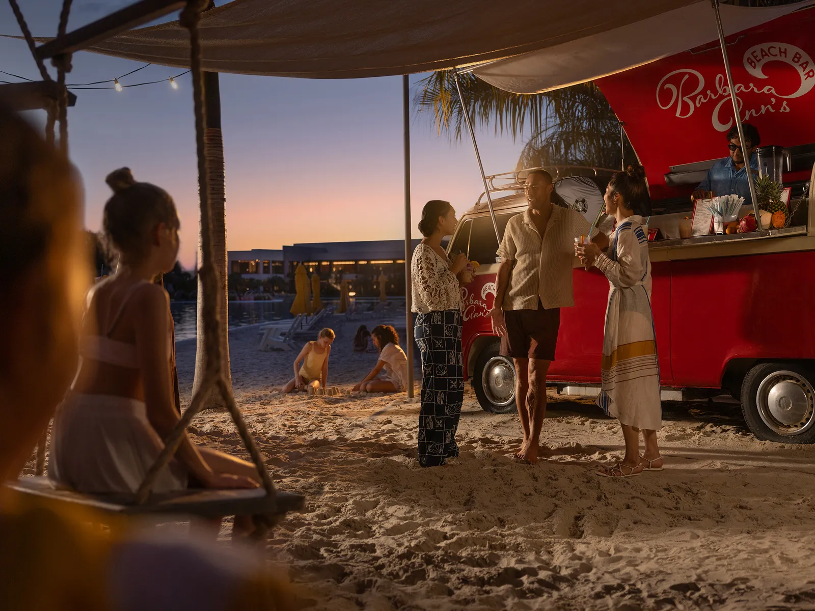 People enjoy drinks by a red beach bar truck on sandy shore at sunset with kids playing nearby.