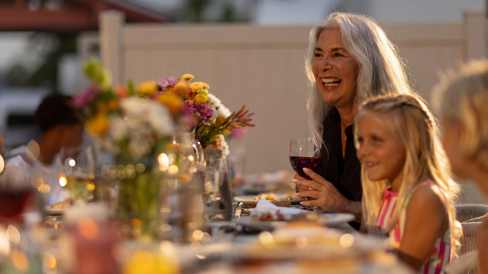 Older woman smiling and holding a glass of red wine at a table with a young girl and others outdoors.