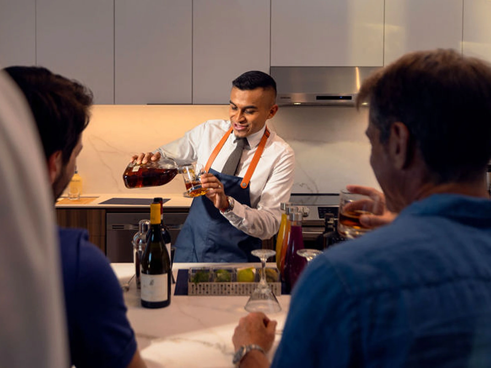 Bartender in apron pouring whiskey into a glass for two men seated at a kitchen bar counter.