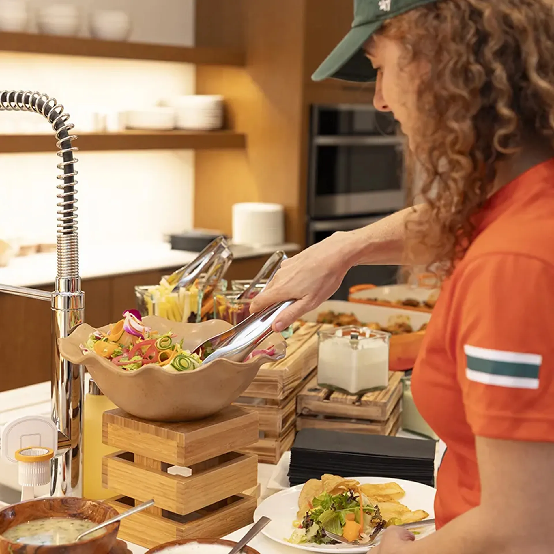 Woman in orange shirt serving salad with tongs from a bowl at a buffet in a kitchen setting.