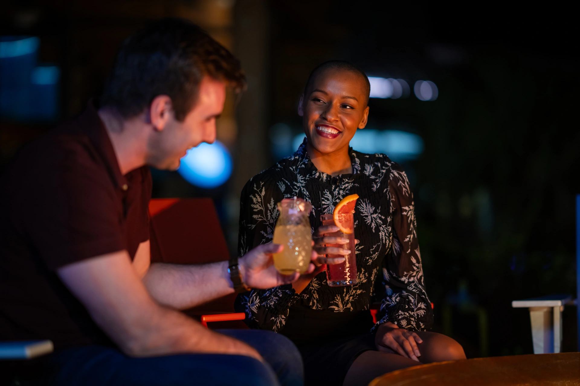 Smiling man and woman sitting outdoors at night, each holding a colorful cocktail drink.