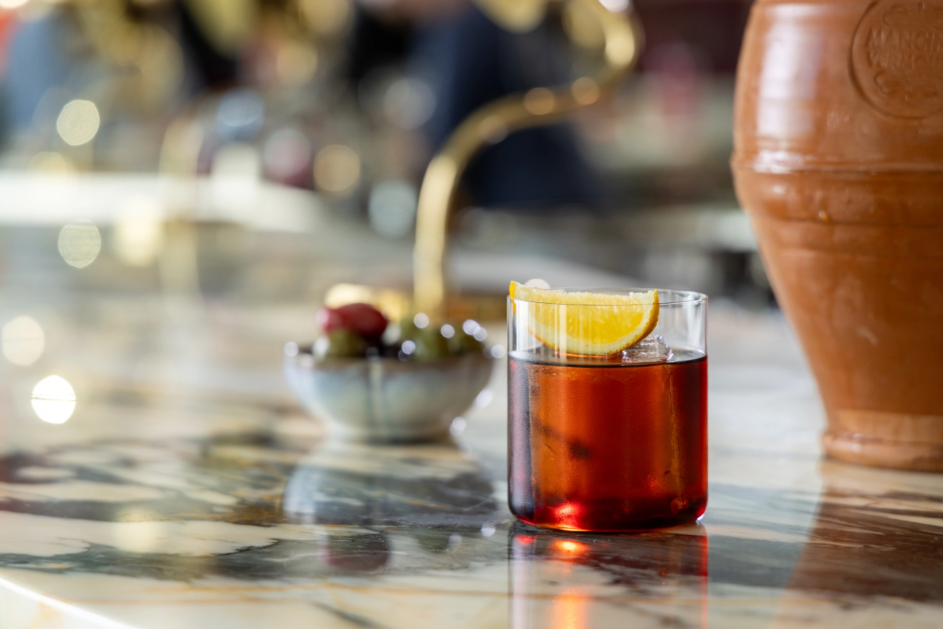 Glass of dark cocktail with an orange slice on a marble counter near a bowl of olives.