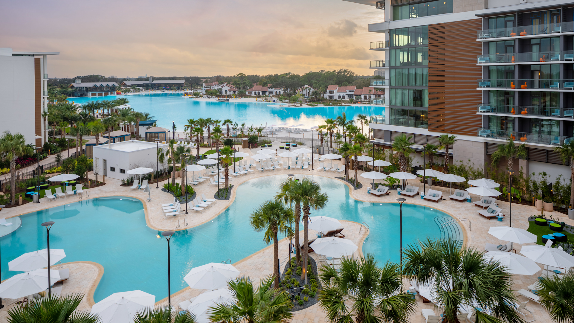 Resort pool area with palm trees, white umbrellas, lounge chairs, and large blue lagoon in the background.