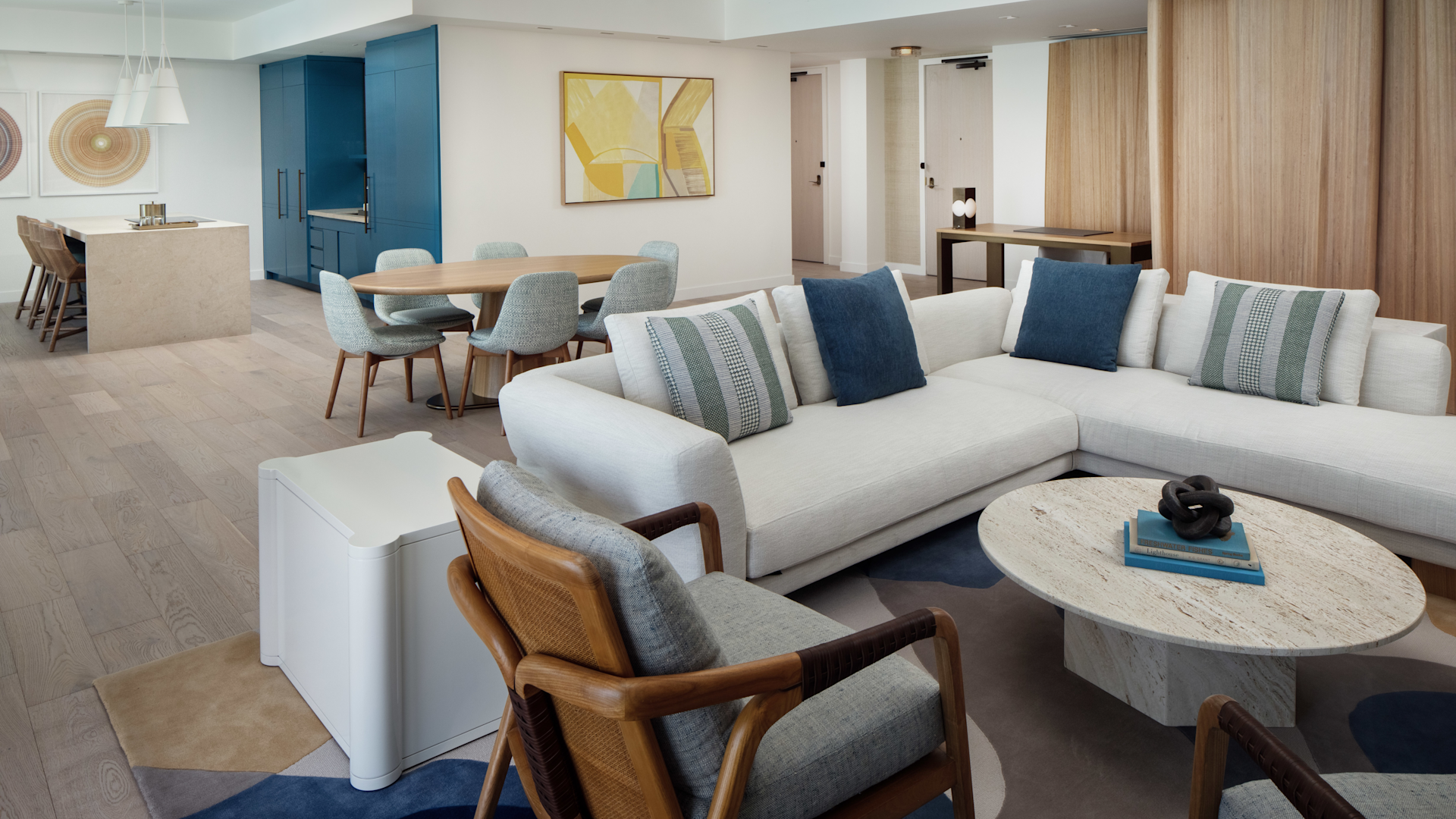 Modern living room with white sectional sofa, wooden chair, round marble coffee table, dining area, and blue cabinet.