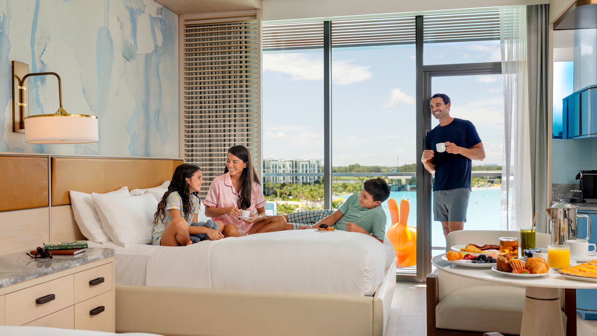 Family relaxing in hotel room with breakfast on table and view of outdoor pool through glass doors.