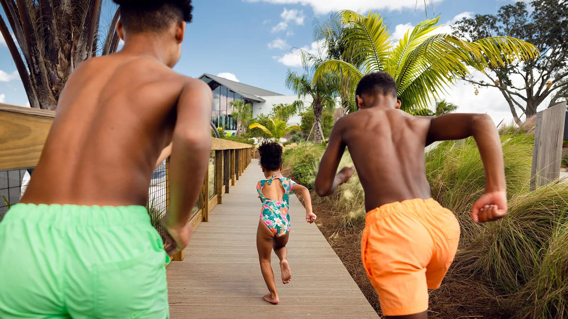 Two boys in swim trunks and a girl in a floral swimsuit running on a boardwalk toward trees and a building.