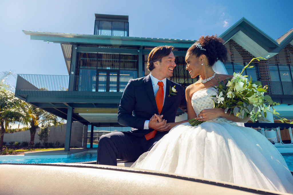 Bride and groom sitting by a pool in front of a modern house, smiling and holding hands.