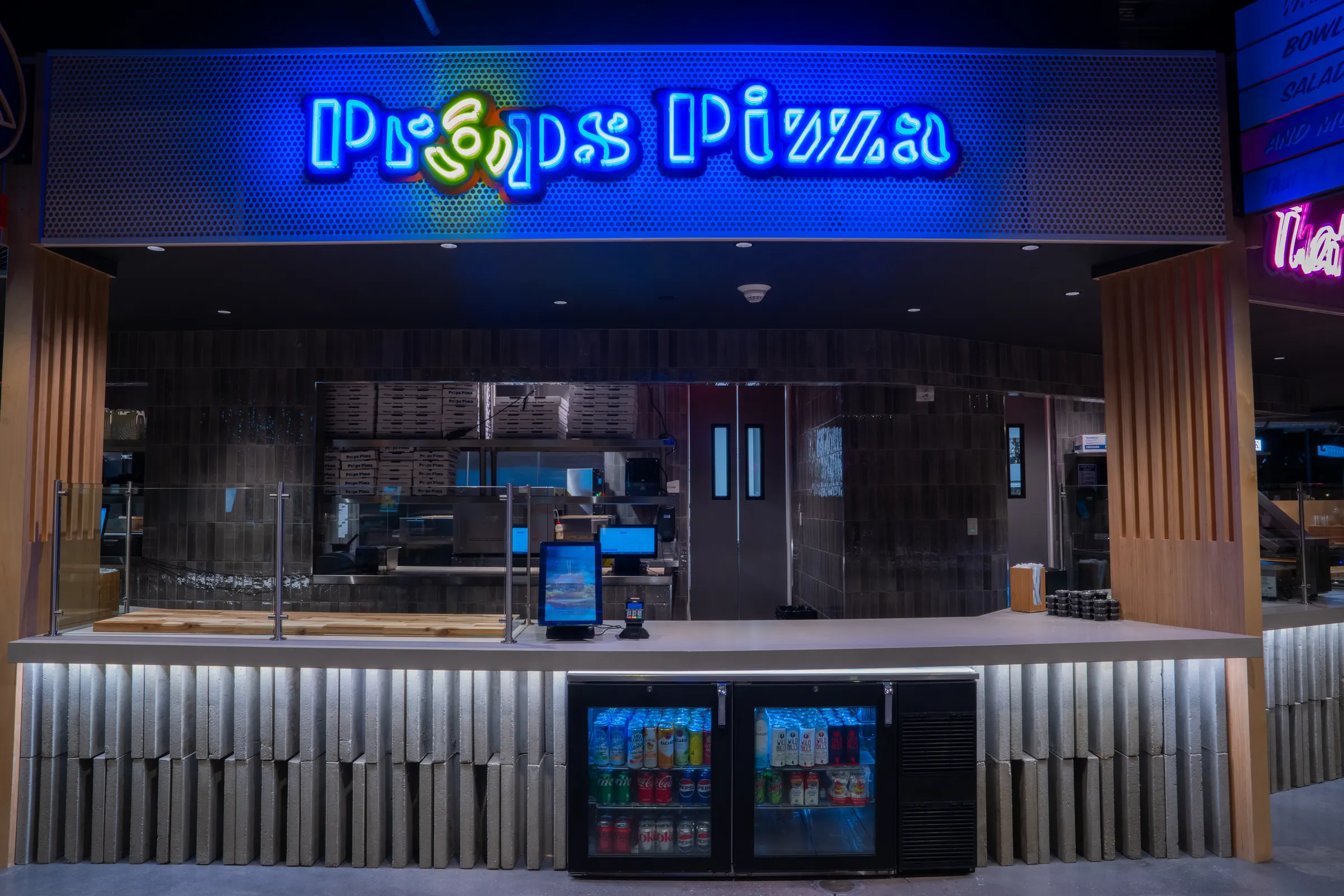 Empty modern pizza counter with a neon sign, drink coolers, and boxed pizzas in background.