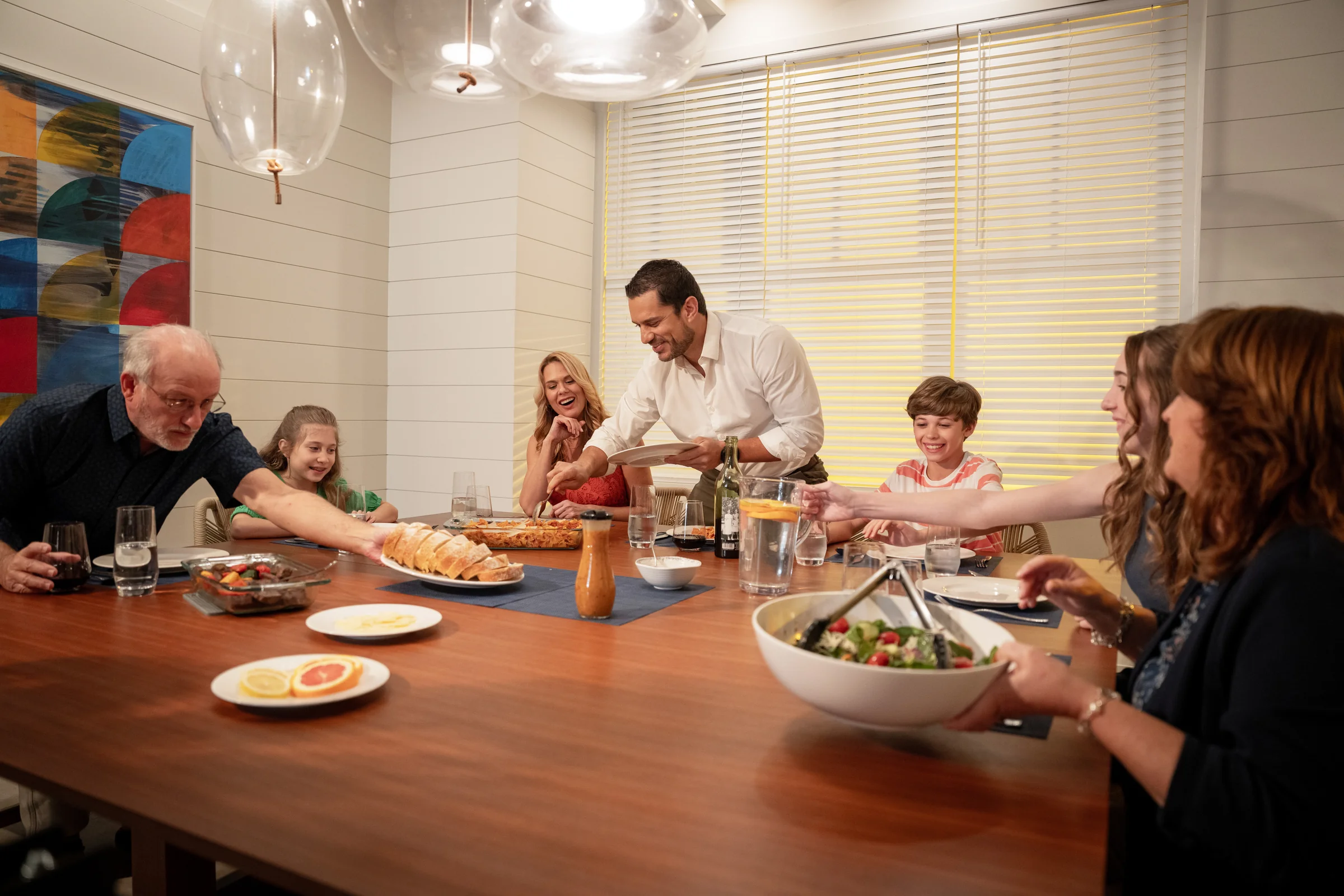 Family of six sharing a meal around a dining table with bread, salad, and drinks in a warm kitchen.