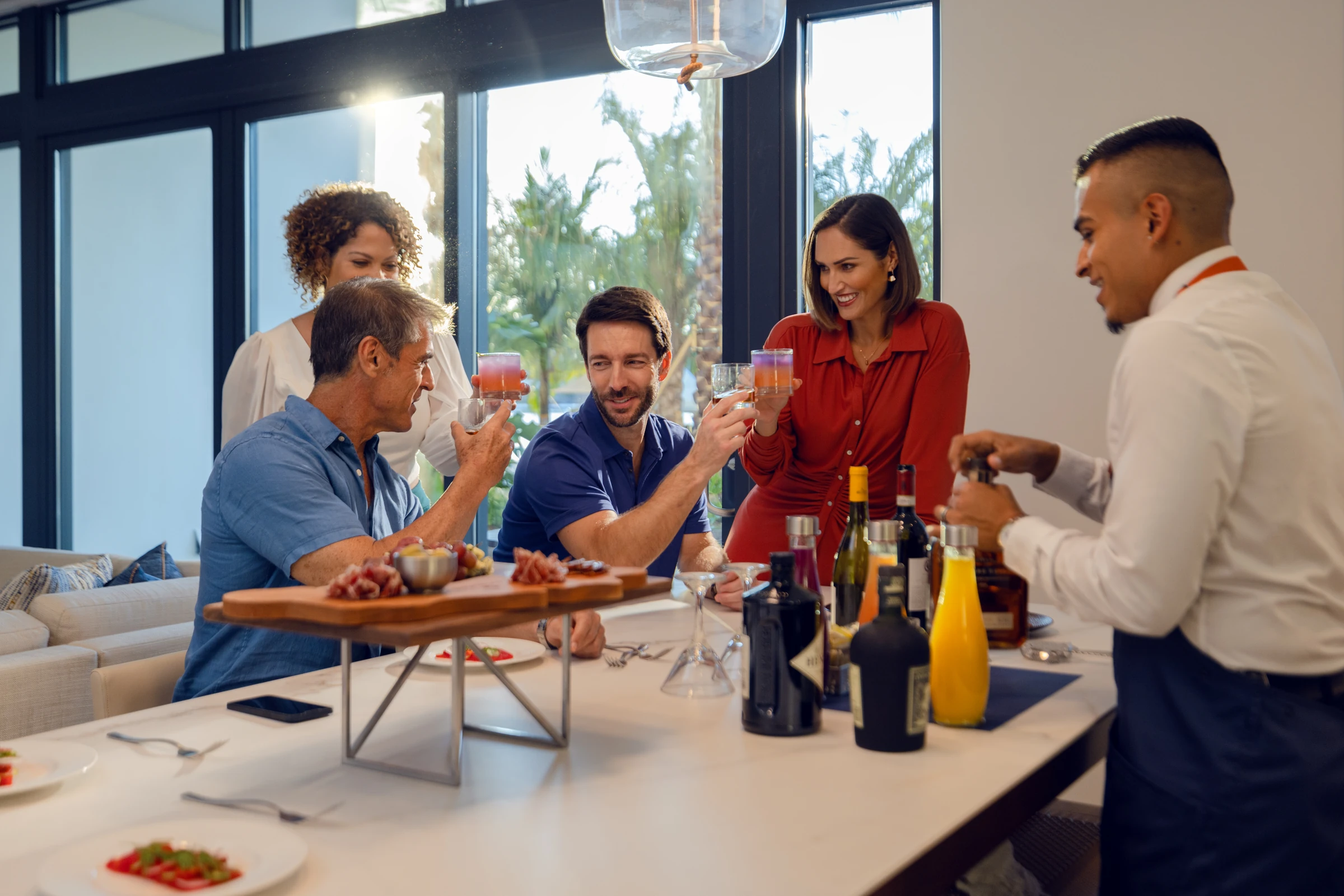 Four friends raising drinks across a table while a bartender opens a bottle nearby.