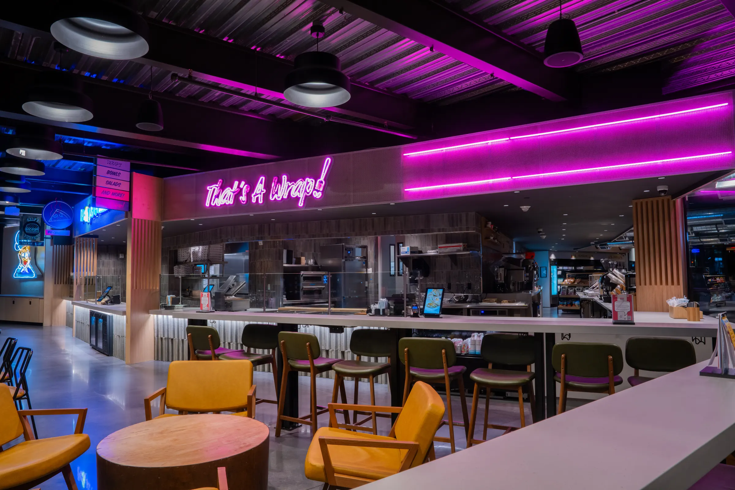 Modern food hall with neon sign saying 'That's A Wrap!' and seating including yellow chairs and stools.