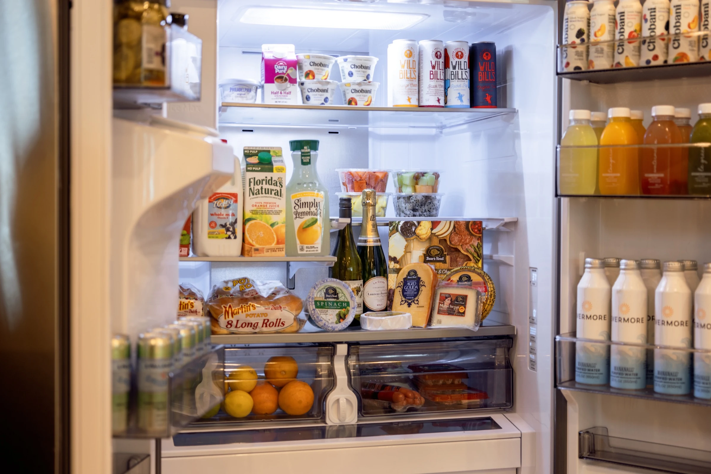 Open fridge stocked with juice, milk, cheese, rolls, fruit cups, wine, and assorted beverages in door shelves.