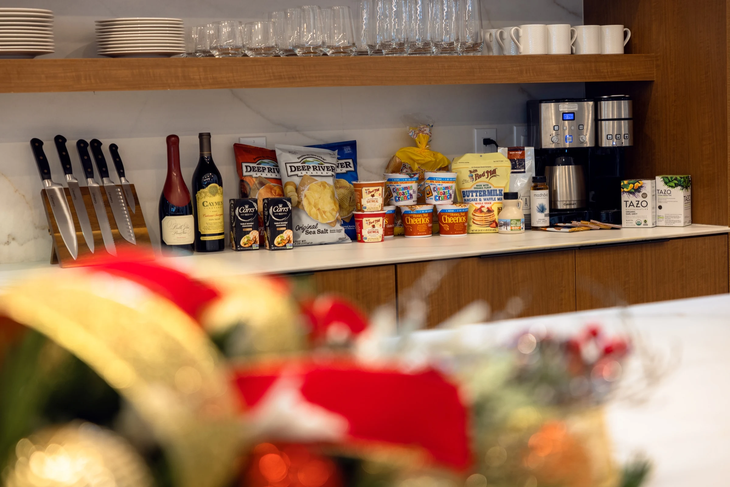 Kitchen counter with knives, wine, snacks, cereal, coffee machine, mugs, and glasses on shelf above.