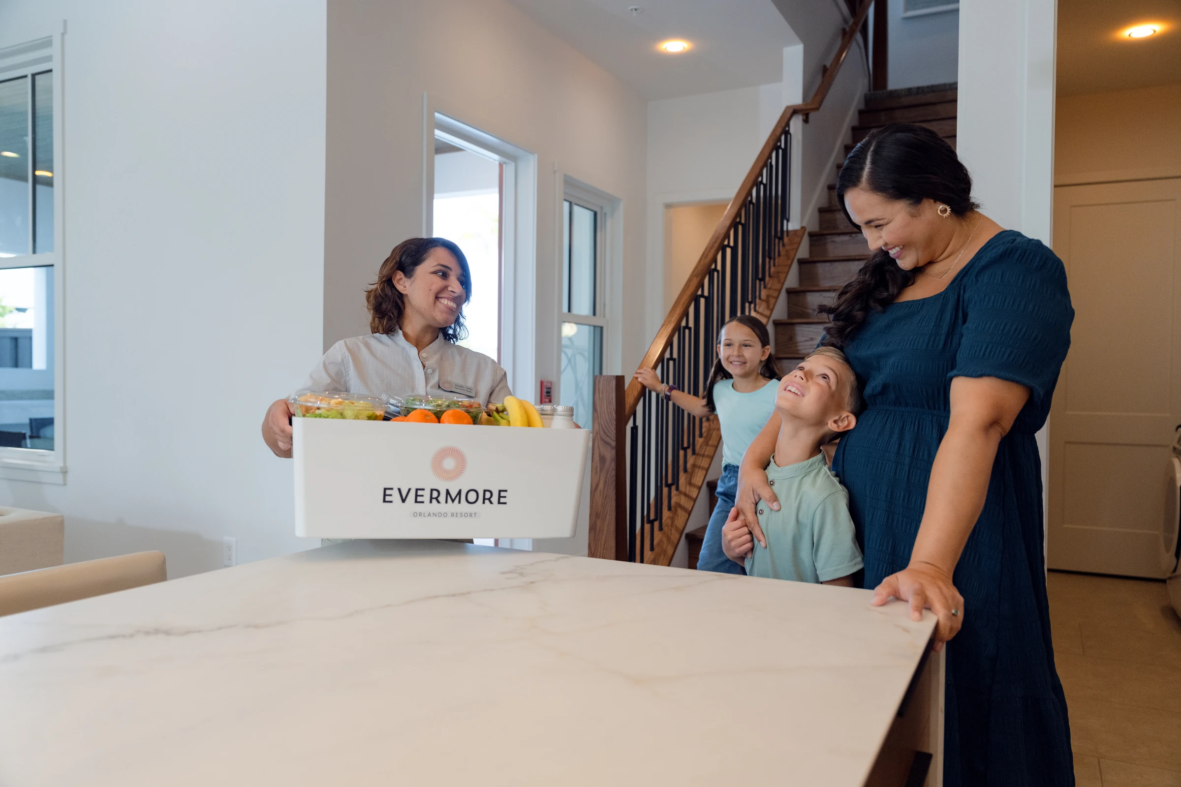 Woman delivering a grocery box labeled Evermore Orlando Resort to a smiling mother with two children.