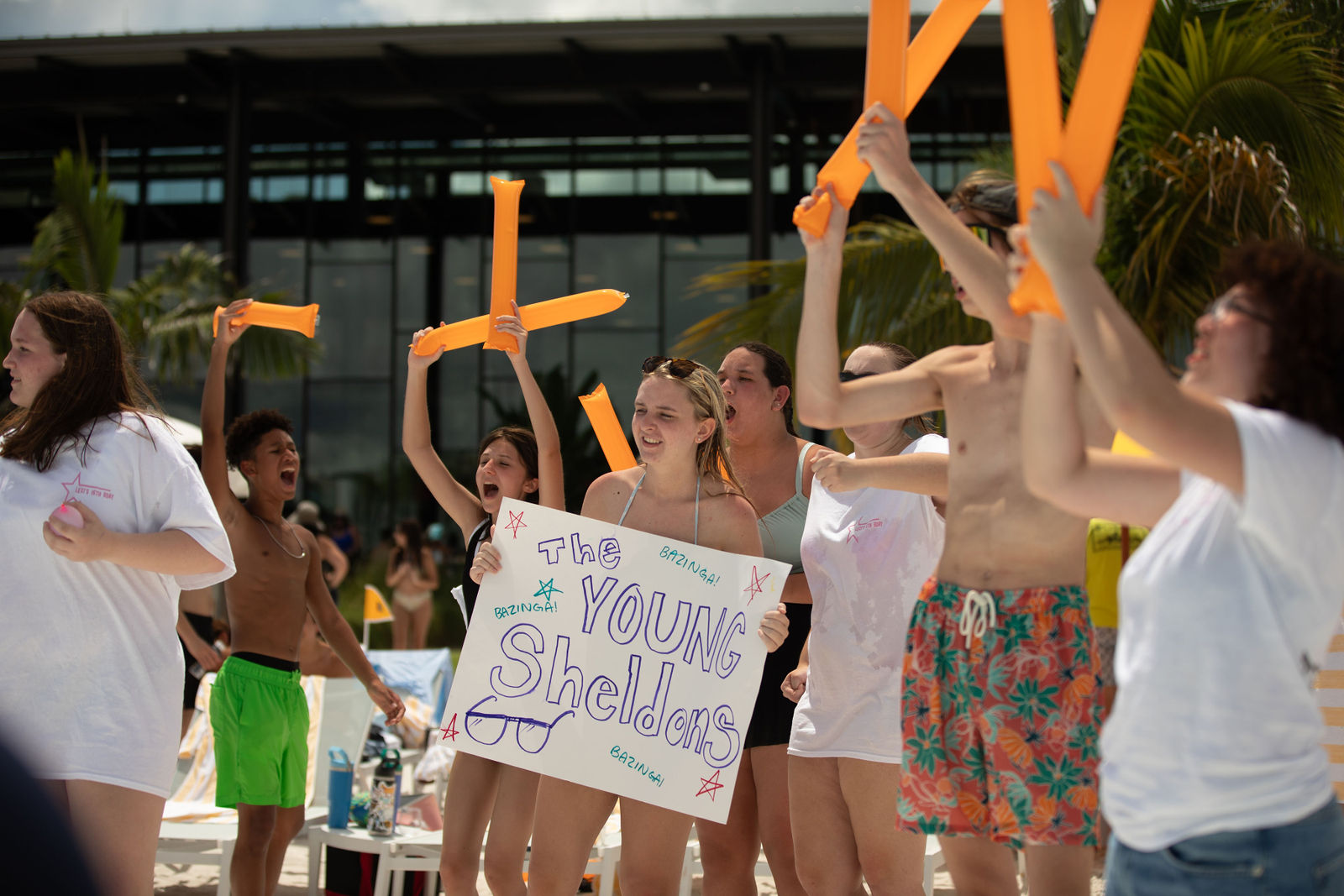 A group of young people cheering and holding orange inflatable sticks and a sign that says The Young Sheldons.