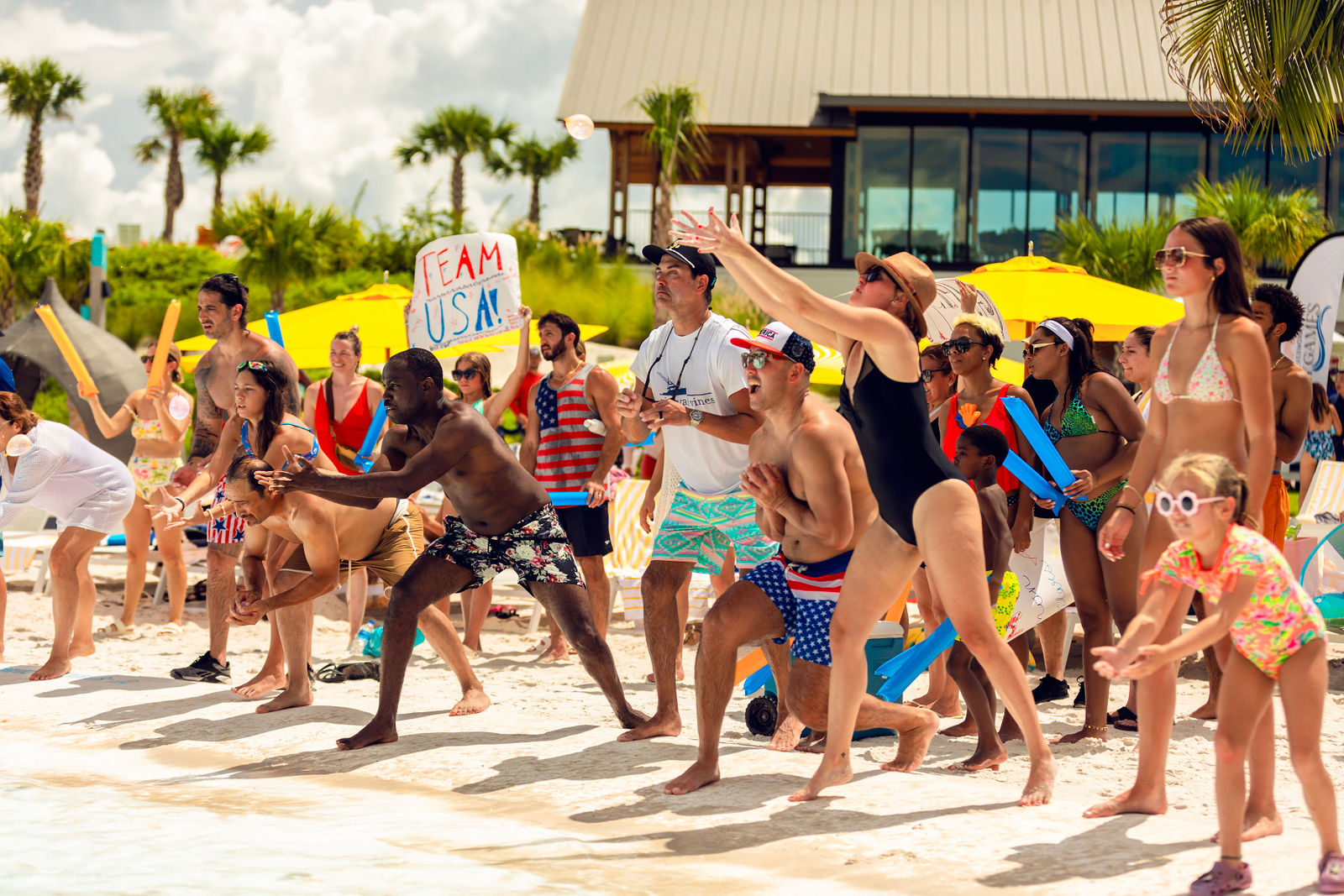 Beachgoers of all ages eagerly catch or reach for water balloons during a sunny outdoor event.