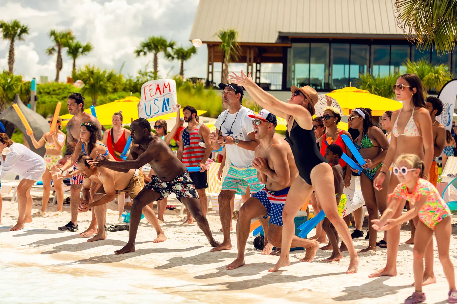 Beachgoers of all ages eagerly catch or reach for water balloons during a sunny outdoor event.